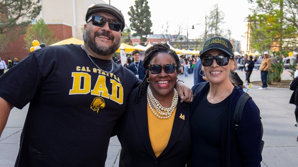 Proud Cal State LA family and community members enjoying a sunny day on campus during a celebration event. Three supporters are smiling and posing together; one wears a “Cal State LA Dad” shirt, one wears a blazer with pearls, and one wears a Cal State LA cap and sunglasses. Tents and other attendees are visible in the background.